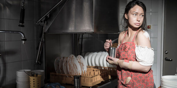 Kvinna med rött förkläde diskar i restaurangkök, ser rädd ut.