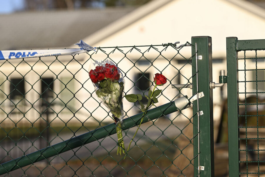 Några röda rosor hänger på ett grönt staket där det också sitter ett polisband. I bakgrunden ett vitt hus.