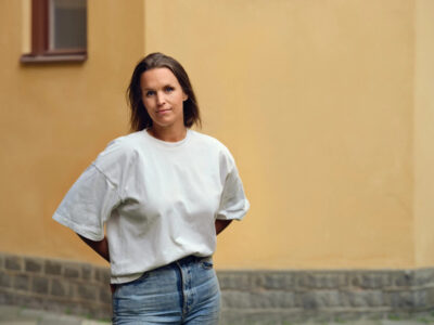 Kvinna i vit t-shirt, framför en husvägg.