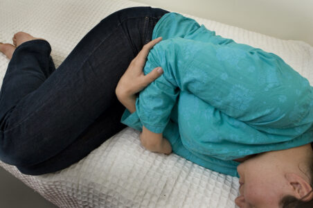 En kvinna ligger i fosterställning och håller sig om magen på en säng.