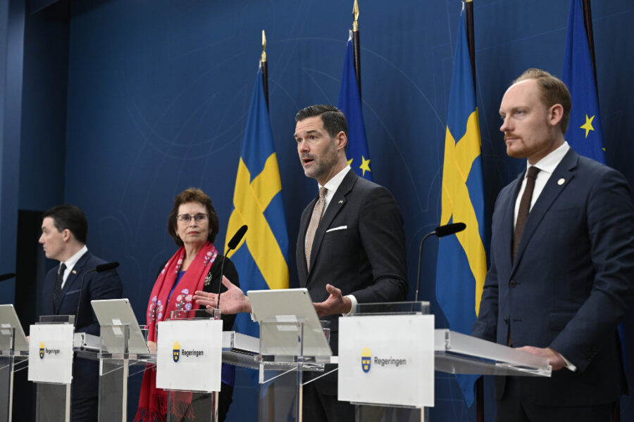 Fyra medlemmar av svenska regeringen intill två svenska flaggor mot blå bakgrund.