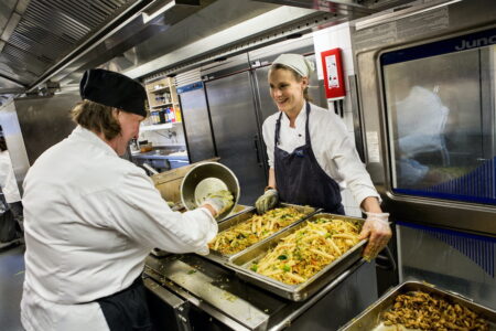 två kvinnor i ett skolkök med metallformar med mat mellan sig