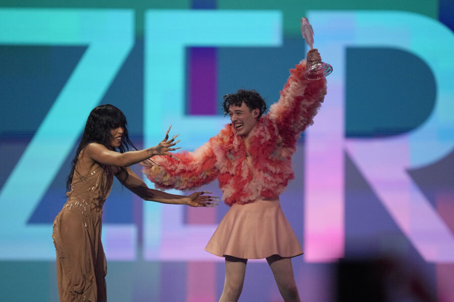 Nemo vinnaren i Eurovision 2024 i rosa kjol och jacka håller upp priset på scen. Bredvid står sångerskan Loreen i en beige klänning