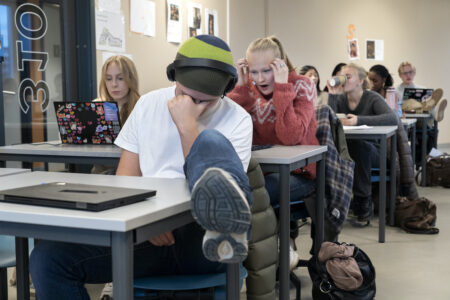 Trötta elever i ett klassrum. En har handen för ansiktet och benet på bänken. En elev dricker ur en pappersmugg en annan håller händerna vid huvudet