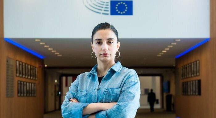 Evin Incir i blå denimskjorta och med armarna i kors. bakom syns EU-flaggan