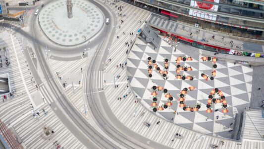 Svartrutigt torg fotat uppifrån där en formation skriver Nej DCA