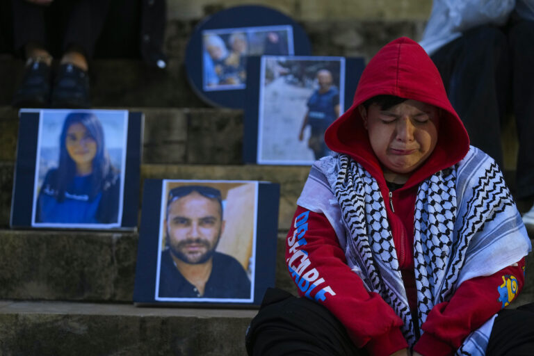 En pojke med röd luvtröja och palestinasjal sitter på en trapp och gråter. Bakom porträttbilder på journalister