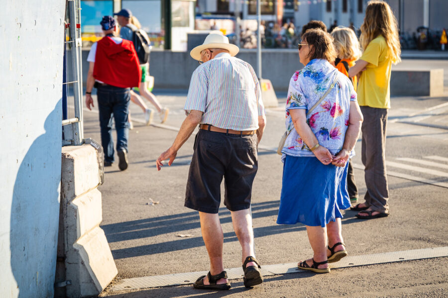 Två äldre personer fotade bakifrån ute på promenad en solig dag.