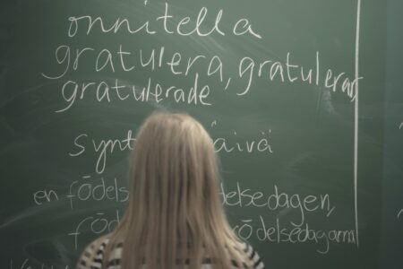 Flicka med långt ljust hår står och skriver på finska och svenska på en grön tavla