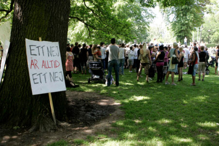 Ett plakat står lutat mot en trädstam, i bakgrunden demonstranter med ryggen mot kameran.