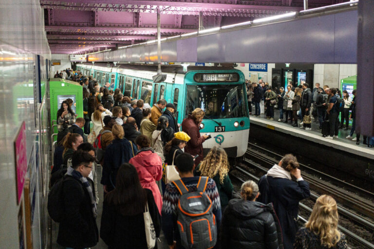 Folk väntar på perrong, turkosafärgat tunnelbanetåg rullar in på stationen.