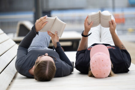 Två unga kvinnor ligger på träbänk och läser böcker.