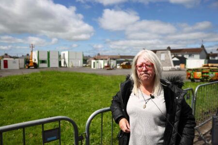 Anna Corrigan står vid stängsel där utgrävning av massgrav inlettts.