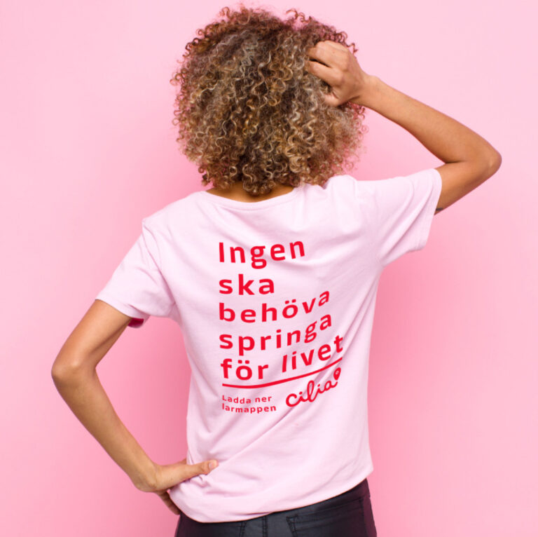 Kvinna med hand i håret fotad bakifrån, iklädd rosa t-shirt.