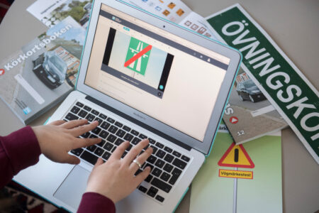 Datorskärm, övningskörningsskylt och handbok för körkort på ett bord.