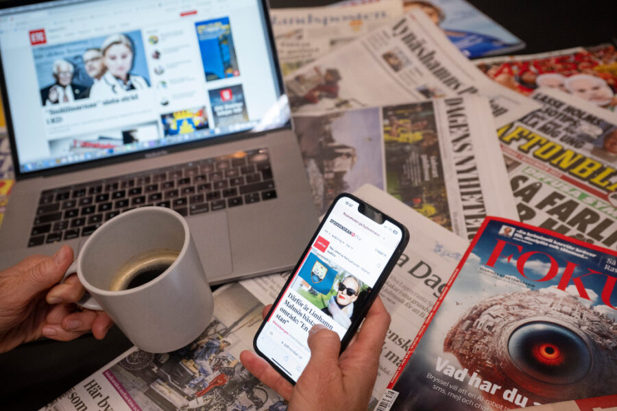 Hand håller i kaffekopp och mobil, framför datorskärm och tidningar.