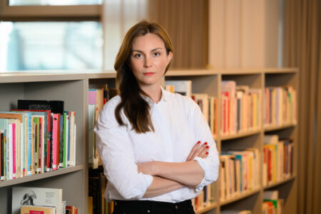 Lina Fridén i vit skjorta med armar i kors framför bokhylla.