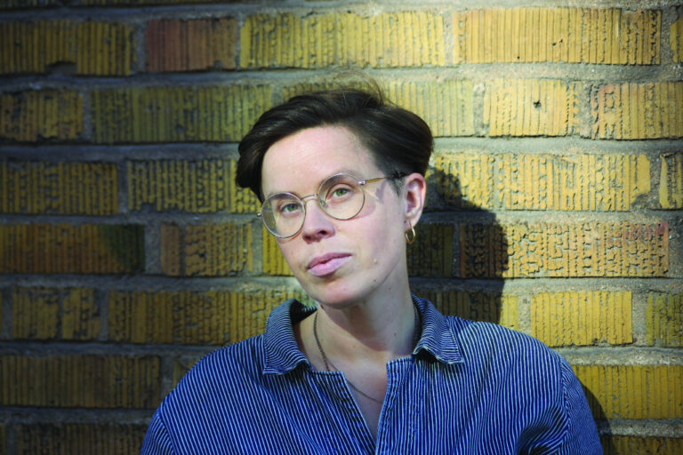 Ylva Emel Karlsson fotad av Josefin Granqvist med en tegelvägg i bakgrunden.