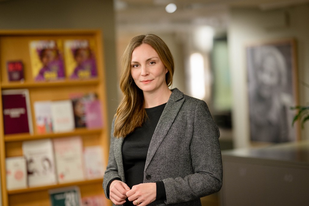 Lina Fridén i kavaj, i bakgrunden en bokhylla och ett stort svartvitt porträtt.