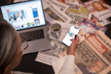 Kvinna med dator och mobil samt tidningar på ett skrivbord..