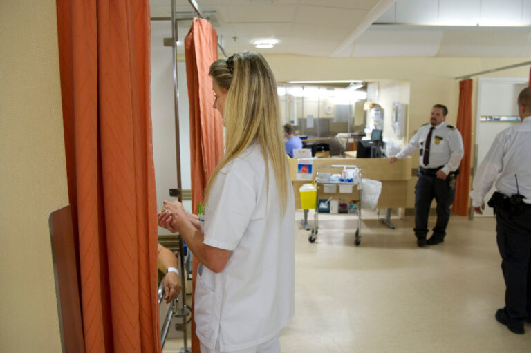 En undersköterska i vitt står vid ett orange draperi, en ordningsvakt längre bort i bakgrunden.