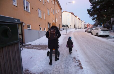 Kvinna drar barnvagn på snötäckt trottoar medan barn går strax framför.